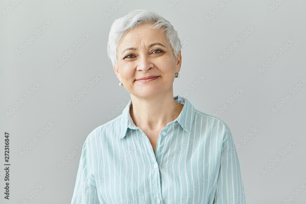Positive human reactions, feelings and emotions. Charming elegant middle aged sixty year old female with short gray hair looking at camera with pleased smile, her eyes full of happiness and joy - obrazy, fototapety, plakaty 