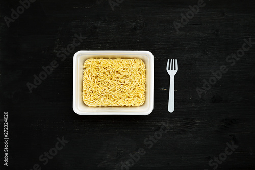 Instant noodles in container with plastic fork on a black wooden table, top view