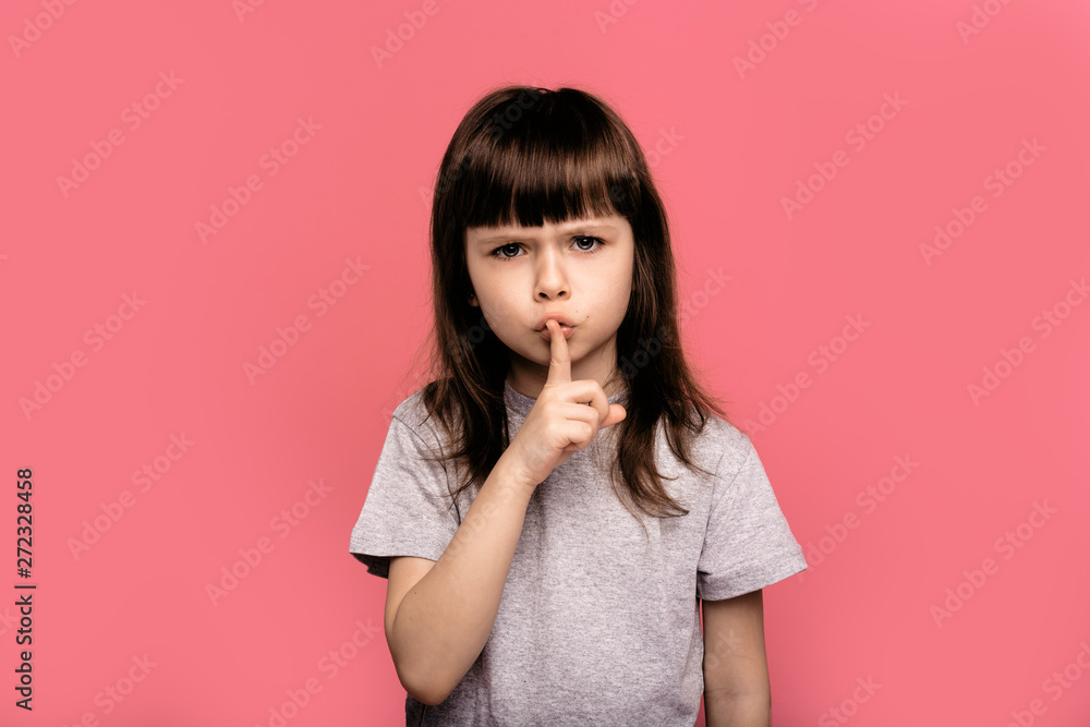 Portrait of girl child keeping finger on her lips and asking to keep quiet, dressed in casual clothes, stands against pink background with blank copy space for your advertisement Portrait of girl child keeping finger on her lips and asking to keep quiet, dressed in casual clothes, stands against pink background with blank copy space for your advertisement