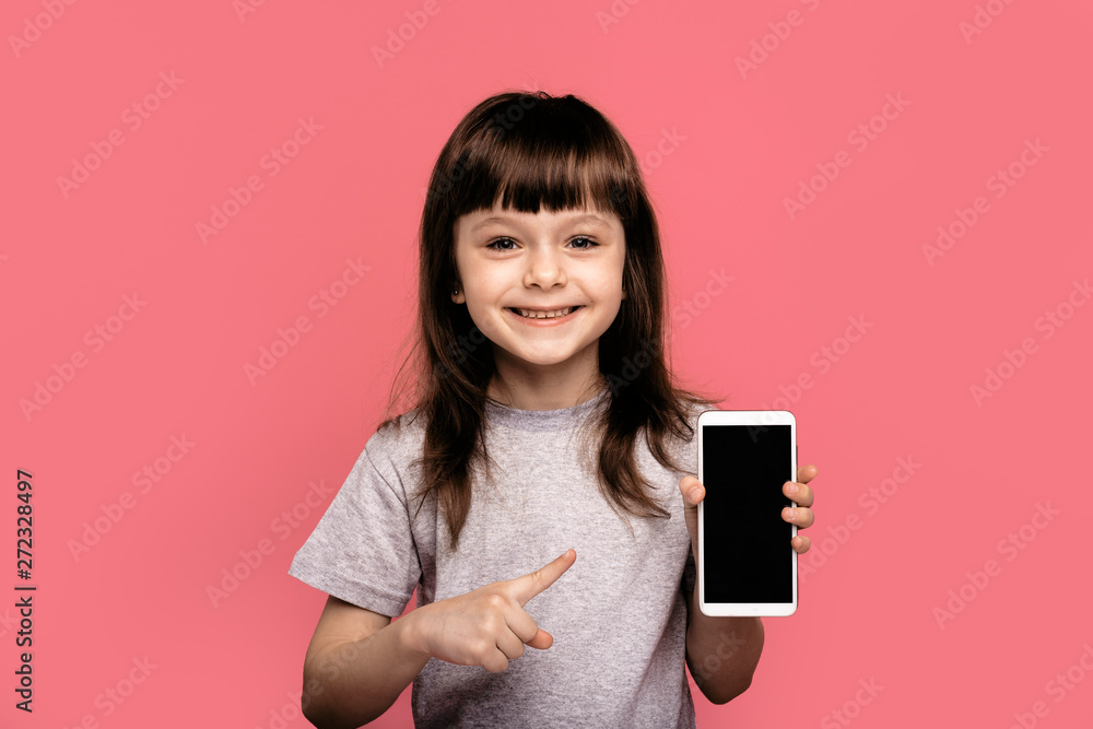 Look at this cell phone! Pleased happy little girl showing blank screen of smart sell phone, modern device, dressed in fashionable t-shirt, isolated over pink background. Technology concept Look at this cell phone! Pleased happy little girl showing blank screen of smart sell phone, modern device, dressed in fashionable t-shirt, isolated over pink background. Technology concept