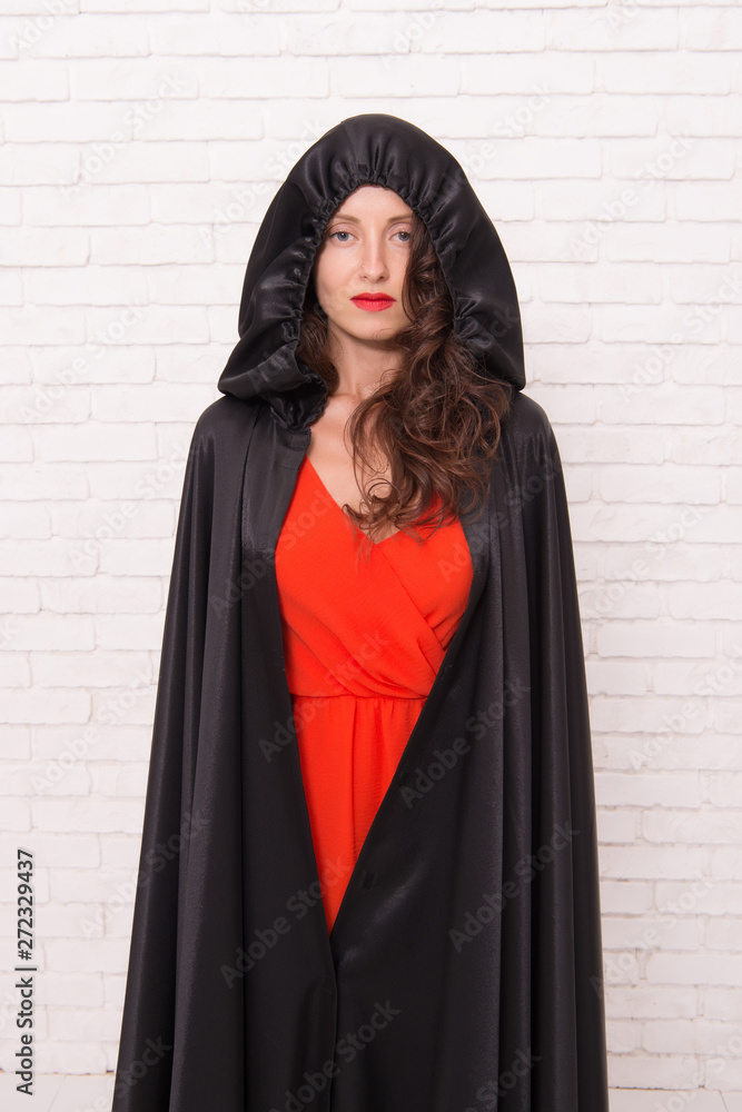 Naklejka premium Her sorcery skills are just far too impressive. Halloween party girl. Sexy girl wearing hooded black cloak on white brickwall. Mysterious girl with classic witch look. A girl changed into a witch