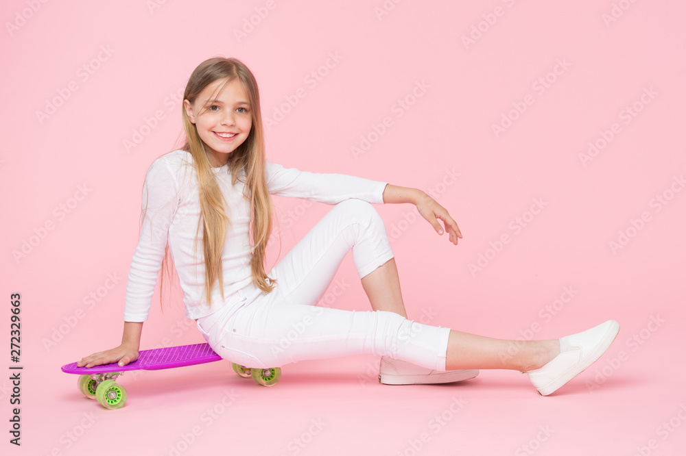 Riding board in style for years to come. Happy little child sitting on board deck on pink background. Cute small skater smiling with violet penny board. Taking pleasure in skateboarding on her board Riding board in style for years to come. Happy little child sitting on board deck on pink background. Cute small skater smiling with violet penny board. Taking pleasure in skateboarding on her board