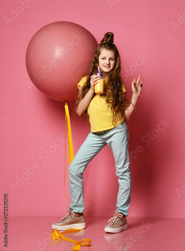 Young teen girl in jeans and yellow t-shirt stepped on big pink balloon yellow tape, drinks yellow juice and wave to us