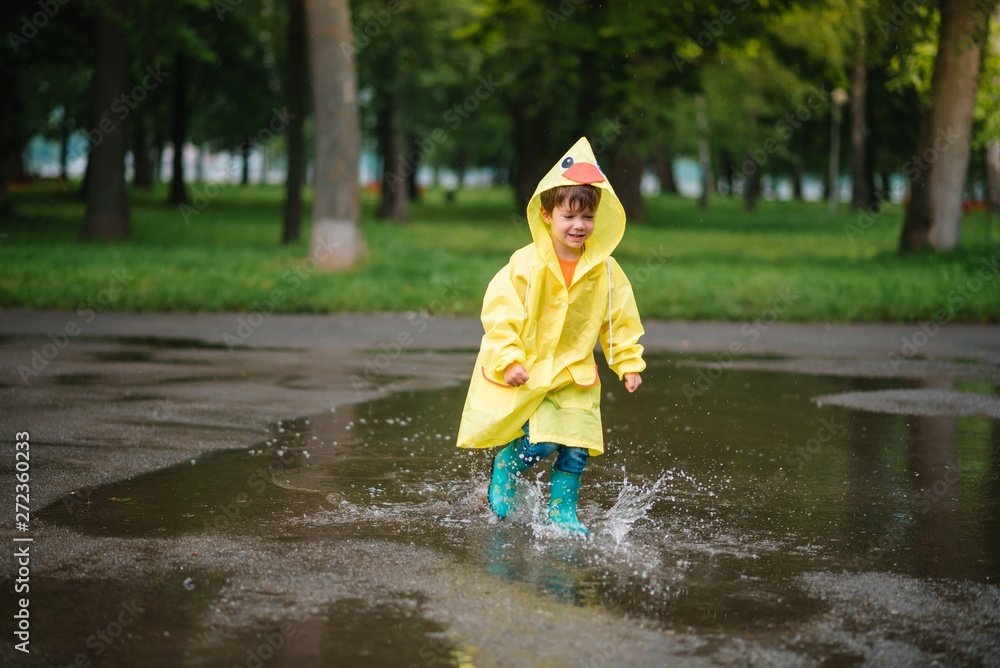 Little boy playing in rainy summer park. Child with umbrella, waterproof coat and boots jumping in puddle and mud in the rain. Kid walking in summer rain Outdoor fun by any weather. happy childhood Little boy playing in rainy summer park. Child with umbrella, waterproof coat and boots jumping in puddle and mud in the rain. Kid walking in summer rain Outdoor fun by any weather. happy childhood