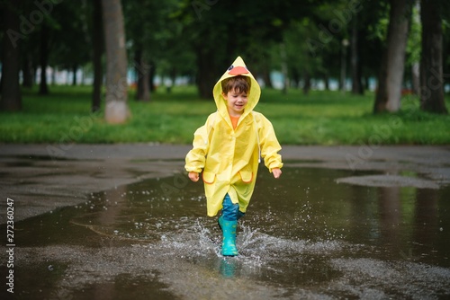 Fototapeta Naklejka Na Ścianę i Meble -  Little boy playing in rainy summer park. Child with umbrella, waterproof coat and boots jumping in puddle and mud in the rain. Kid walking in summer rain Outdoor fun by any weather. happy childhood