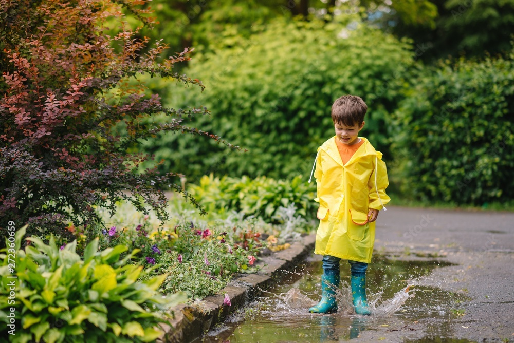 Little boy playing in rainy summer park. Child with umbrella, waterproof coat and boots jumping in puddle and mud in the rain. Kid walking in summer rain Outdoor fun by any weather. happy childhood