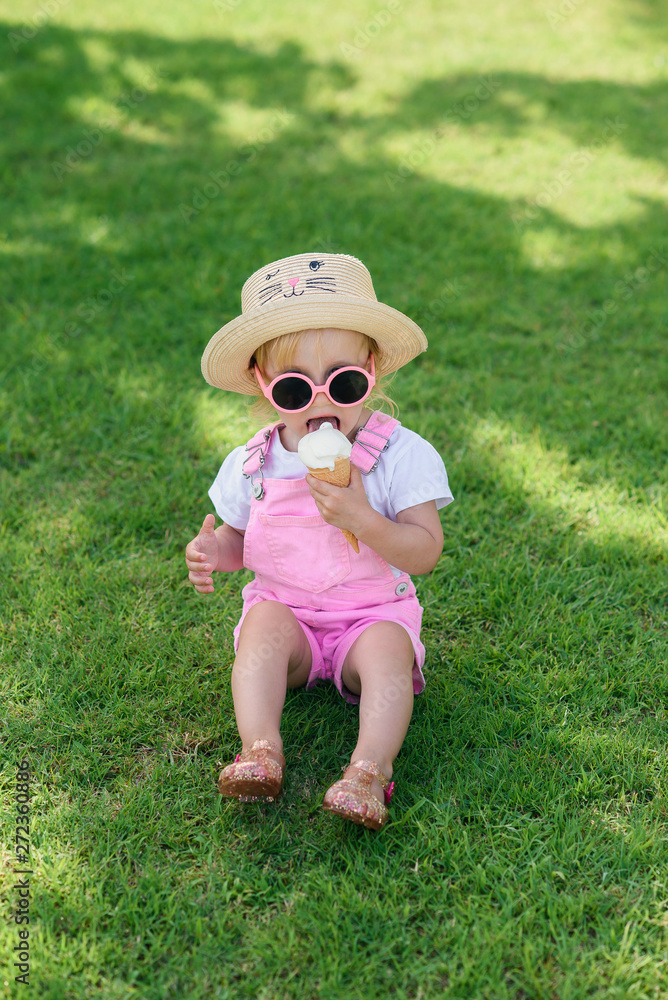 Happy baby girl dressed pink summer clothes, yellow hat and pink sunglasses sits on a green lawn and eats white ice cream in a sunny garden. Happy baby girl dressed pink summer clothes, yellow hat and pink sunglasses sits on a green lawn and eats white ice cream in a sunny garden.