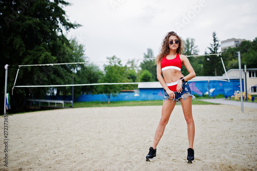 Sexy curly model girl in red top, jeans denim shorts, cup and sunglasses posed at beach volleyball field. © AS Photo Family
