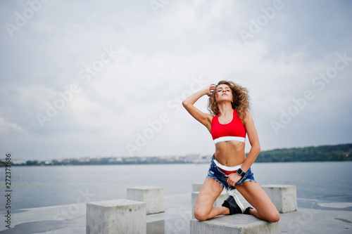 Sexy curly model girl in red top, jeans denim shorts and sneakers posed at stone cubes against cloudy sky and lake.