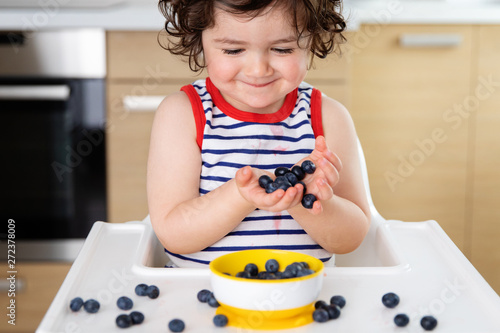 Smiling toddler in high chair holding blueberries in hands