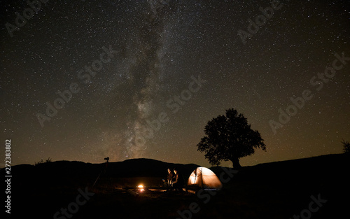 Two men sitting on big logs between tourist tent and tripod camera, having a rest at burning bonfire under night sky full of stars and Milky Way constellation. Night camping, tourism and photography.