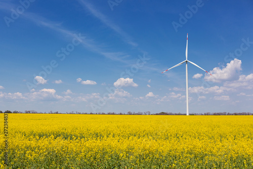in the yellow canola field stands a white turbogenerator, against the sky with clouds, the concept © aneduard