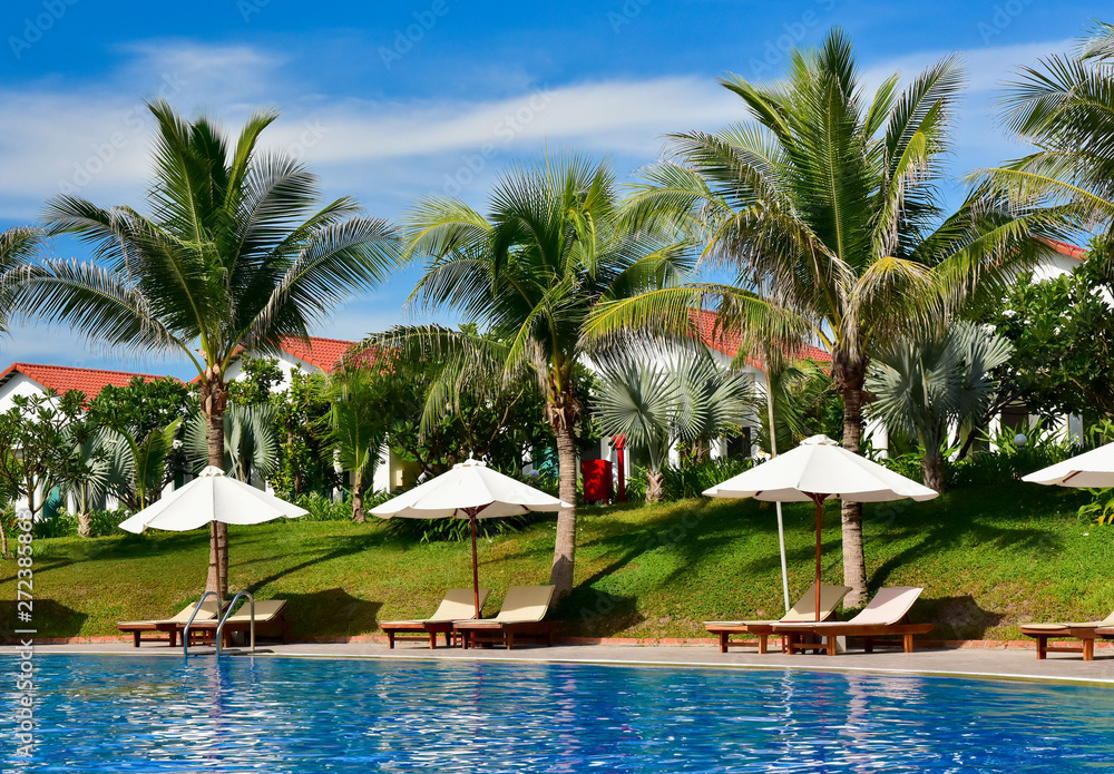 empty deck chairs with sun umbrellas next to the pool. The territory of a modern hotel in Asia. bungalow roofs behind palm trees