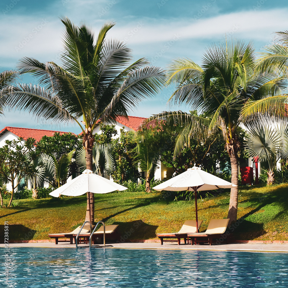 empty deck chairs with sun umbrellas next to the pool. The territory of a modern hotel in Asia. bungalow roofs behind palm trees. vintage toning