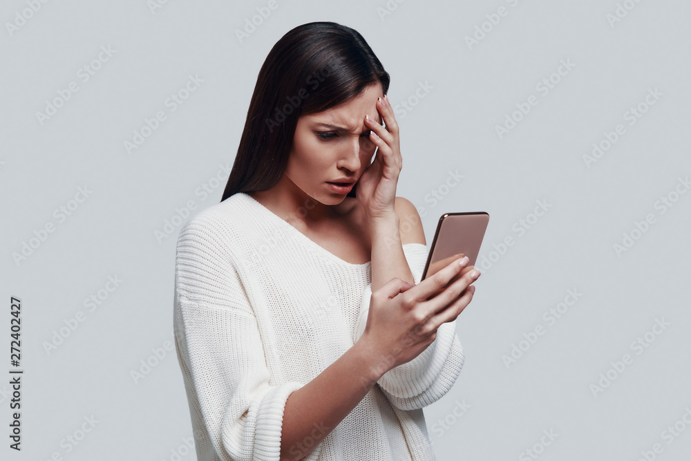 So upset! Confused young woman using smart phone while standing against grey background