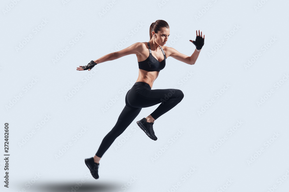 Full of energy. Full length of attractive young woman in sports clothing jumping while hovering against grey background Full of energy. Full length of attractive young woman in sports clothing jumping while hovering against grey background