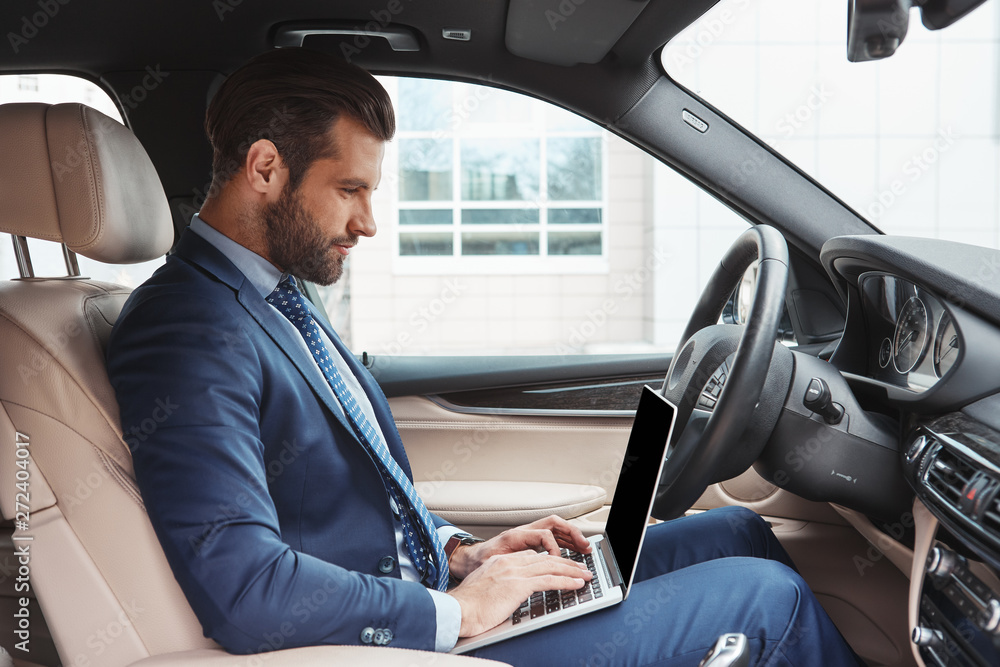 He is available from everywhere. Side view of successful and young bearded businessman working on laptop while sitting in his car He is available from everywhere. Side view of successful and young bearded businessman working on laptop while sitting in his car