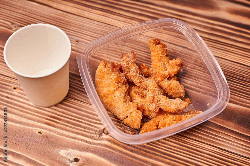 Chicken strips in a plastic container with a paper Cup on a wooden table. Chicken strips in a plastic container with a paper Cup on a wooden table.