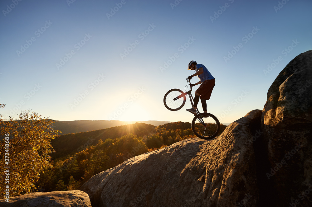 Fearless cyclist standing on back wheel on trial bicycle. Professional sportsman rider balancing on the edge of big boulder on the top of mountain at sunset. Concept of extreme sport Fearless cyclist standing on back wheel on trial bicycle. Professional sportsman rider balancing on the edge of big boulder on the top of mountain at sunset. Concept of extreme sport