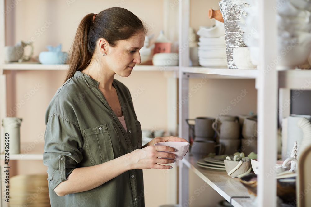 Side view portrait of contemporary female potter putting handmade bowl on shelf in studio, copy space