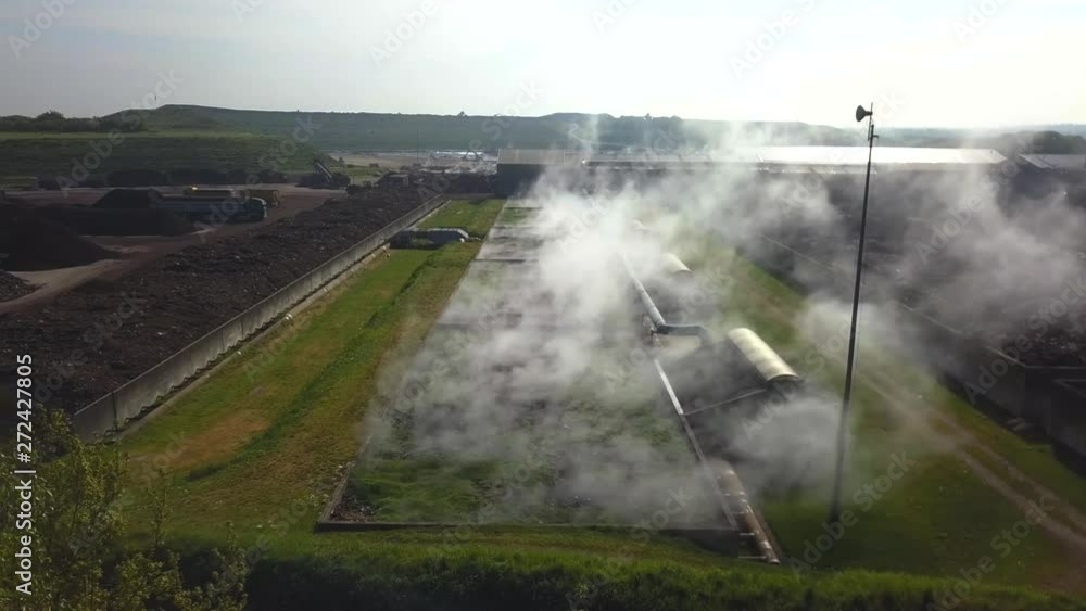 Forward drone shot, from behind some trees, of an industrial compost biofilter, with steam going up, surrounded by dunghills and green fields Forward drone shot, from behind some trees, of an industrial compost biofilter, with steam going up, surrounded by dunghills and green fields