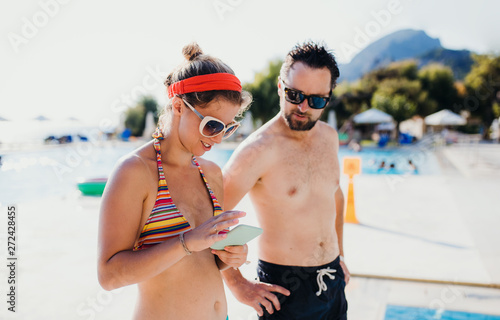 A couple in swimsuit by the swimming pool on summer holiday, using smartphone.