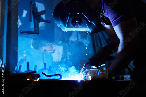 Busy factory worker in protective mask using torch while welding wrought iron in dark workshop  blue light from welding process