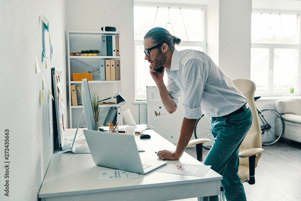 Checking every detail. Good looking young man in shirt talking on the smart phone and smiling while working in the office Checking every detail. Good looking young man in shirt talking on the smart phone and smiling while working in the office