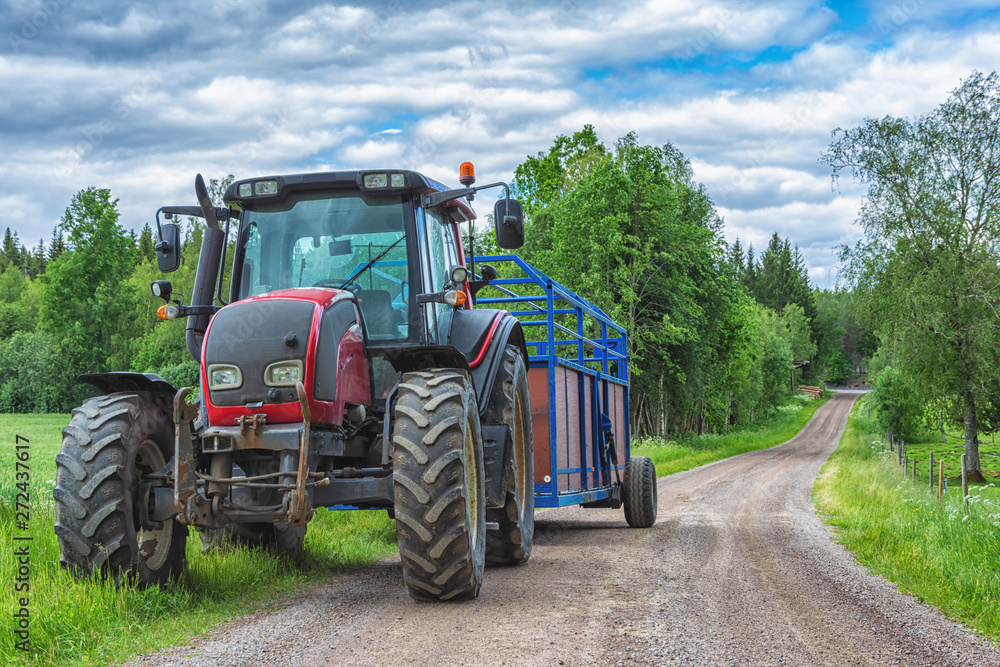 Tractor with trailer on the country side road with green trees and cloudy sky