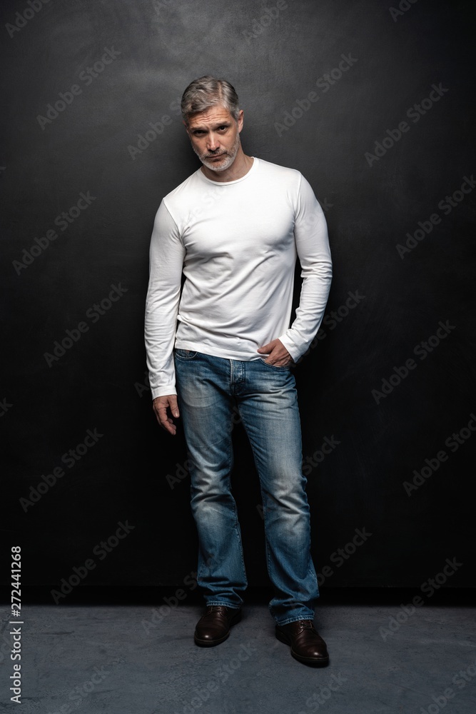 Obraz premium Full body portrait of middle-aged good looking man in white t-shirt posing in front of a black background with copy space.