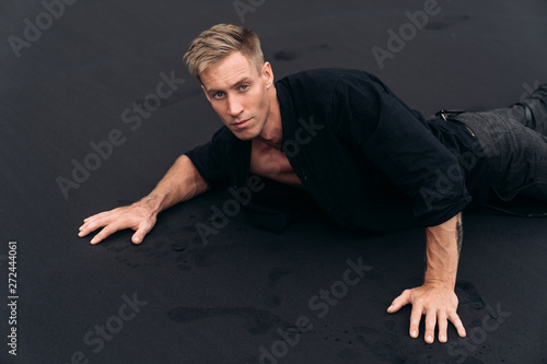 Portrait of handsome blue-eyed man in black shirt lying on black sand beach