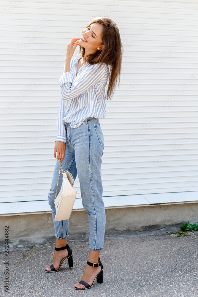 Young stylish woman wearing blue striped shirt, blue cropped denim jeans,  black high heel sandals and beige handbag posing outdoors against white  street wall. Trendy casual outfit. Street fashion. Stock Photo