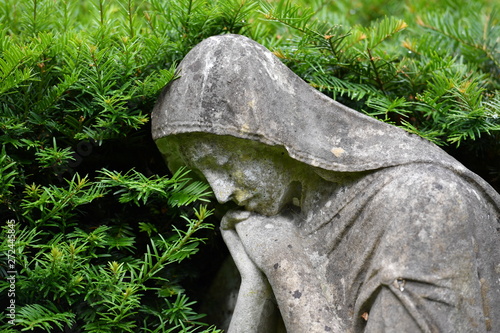A weathered statue of a sad angel that is surrounded with by branches on a christian cemetery in Berlin-Germany.