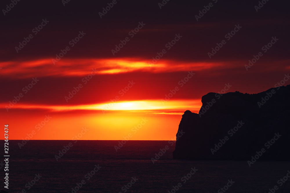 Dramatic ocean sunset scene with giant big sun setting behind a rock coastline silhouette an colorful sky glow. Port Andratx, Mallorca, Spain