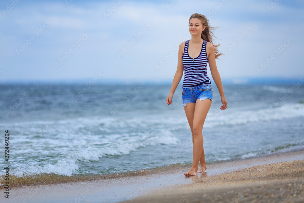 beautiful happy girl walking on the beach in a striped t-shirt and denim shorts, hair fluttering in the wind beautiful happy girl walking on the beach in a striped t-shirt and denim shorts, hair fluttering in the wind