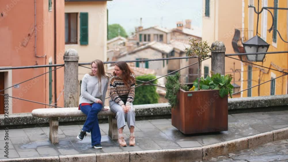 Two young women are sitting on the bench in outdoor. Flowerbed is near Two young women are sitting on the bench in outdoor. Flowerbed is near