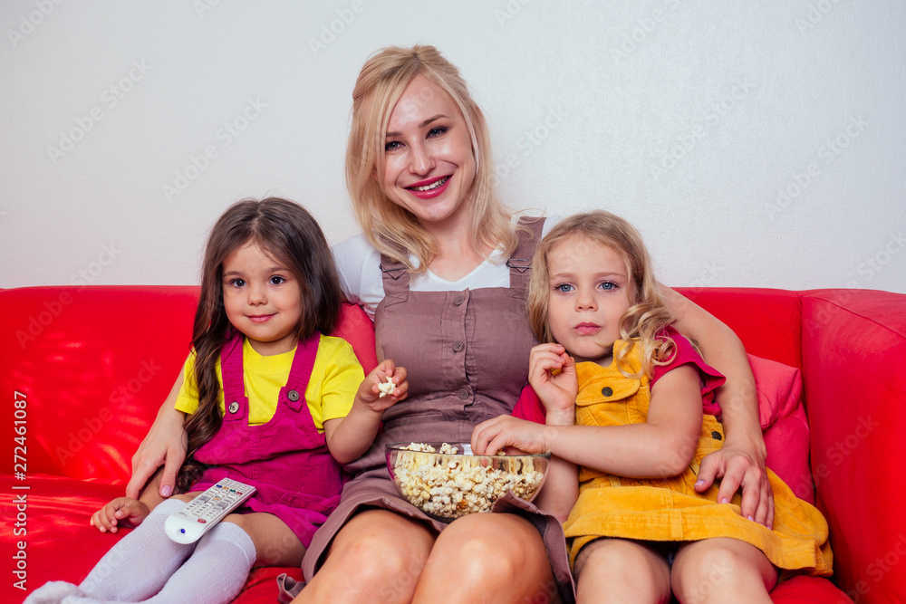 Portrait of happy two daughter and mother watching cartoons with popcorn on a red couch Portrait of happy two daughter and mother watching cartoons with popcorn on a red couch