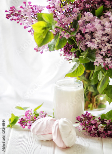 The glass of milk served with handmade marshmallows and a bouquet of a lilac on white table. Breakfast concept. Selective focus. 