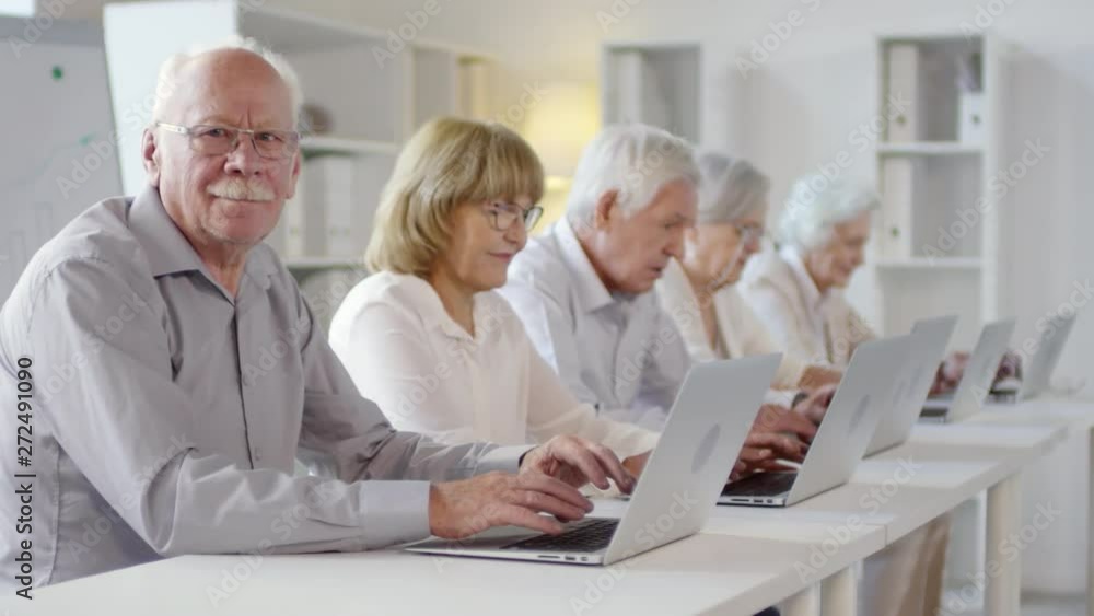 Tracking shot of smiling elderly man with grey hair and mustache looking at camera during computer class for seniors. Old people sitting at desks and typing on laptops Tracking shot of smiling elderly man with grey hair and mustache looking at camera during computer class for seniors. Old people sitting at desks and typing on laptops