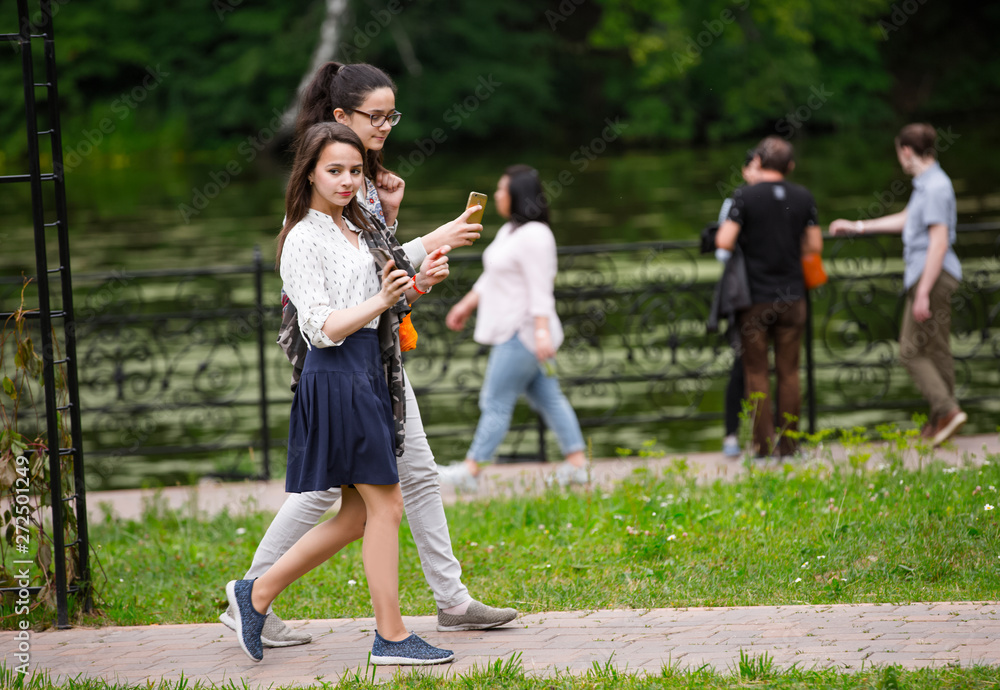full length two friends having walk in a park in Russian traditional park in Moscow. young girls communicating and using mobile phones by nature pool. women travelers browse map app full length two friends having walk in a park in Russian traditional park in Moscow. young girls communicating and using mobile phones by nature pool. women travelers browse map app