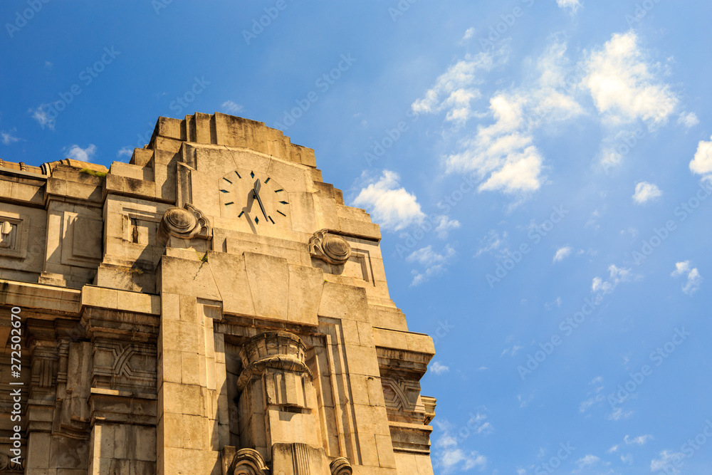 Round clock on top of historical stone building of the main train station (Milano Centrale) in Milan, Italy. Sunny day with a few fluffy clouds in a blue sky.