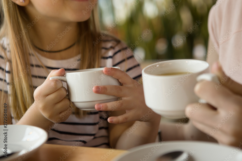 Selective focus of cups of green tea in hands of little girl and woman in cafe. Daughter and mother drinking cappuccino and coffee during lunch, talking and smiling. Concept of coffee and enjoyment. Selective focus of cups of green tea in hands of little girl and woman in cafe. Daughter and mother drinking cappuccino and coffee during lunch, talking and smiling. Concept of coffee and enjoyment.