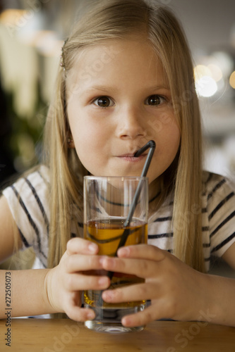 Portrait of funny girl drinking fresh juice in cafe. Little long haired girl sitting at table  keeping glass in hands  looking at camera and posing. Concept of happiness and lunch.