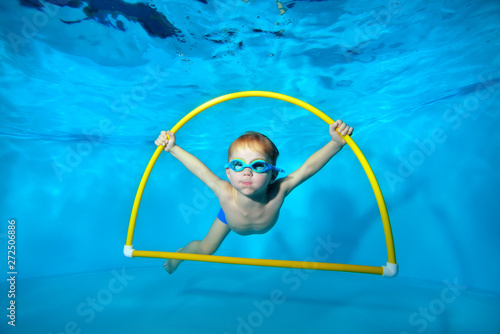 A little sports boy is engaged in swimming in the pool, underwater at the bottom, posing with a yellow Hoop in his hands and looking at the camera. Underwater photography. Horizontal orientation photo