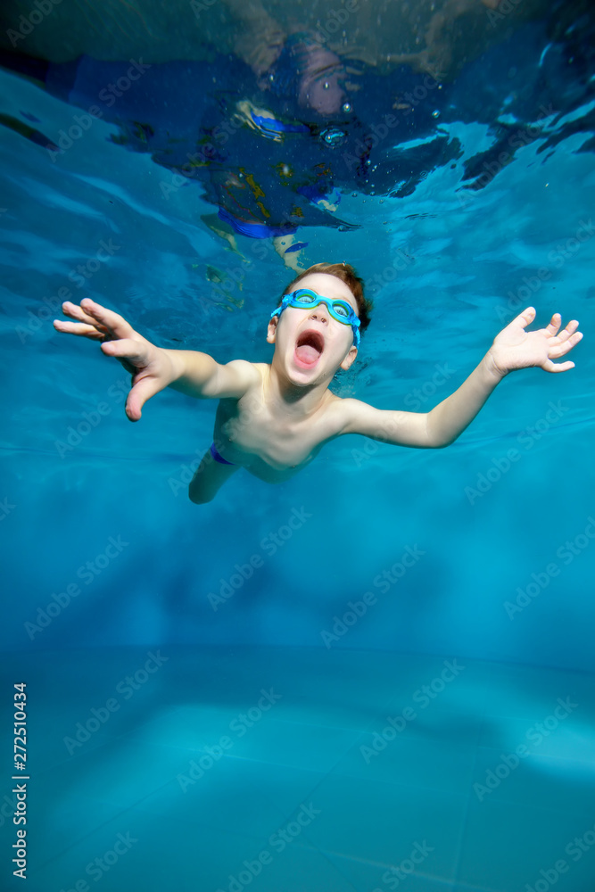 Laughing sports little boy swimming underwater, near the surface of the water. Arms outstretched like a bird. Posing for the camera with his eyes open. Delight. Photo under water. Vertical view