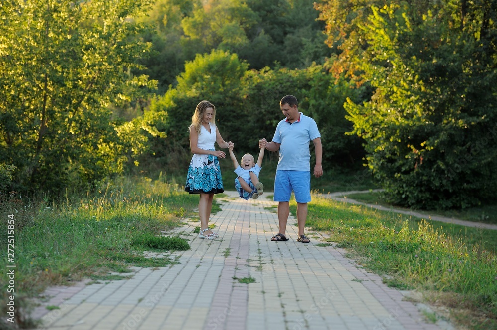 Fototapeta premium Parents swing the little boy in their arms like a swing and walk along the path in the Park at the sunset of a summer day. The concept of family happiness. Shut. Horizontal view