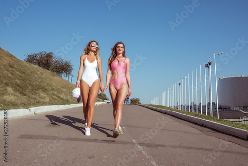 Fototapeta Naklejka Na Ścianę i Meble -  Two girlfriends of the girl in bathing suits, walk in city in the summer, lifestyle fashion, modern trend, tanned figure fitness, walk, sports walking. The concept of a weekend holiday.