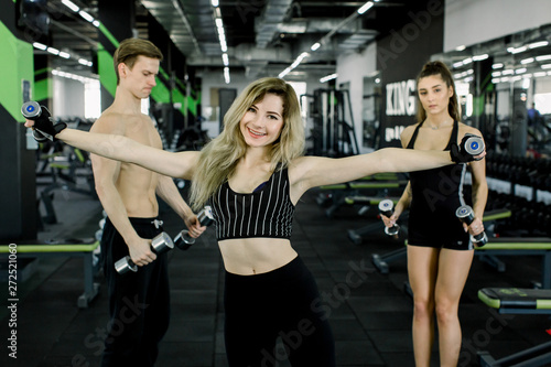 Young people pumping up muscles with dumbbells. Pretty blond fitness girl exercising with barbell in gym together with her friends, woman and man in sport wear