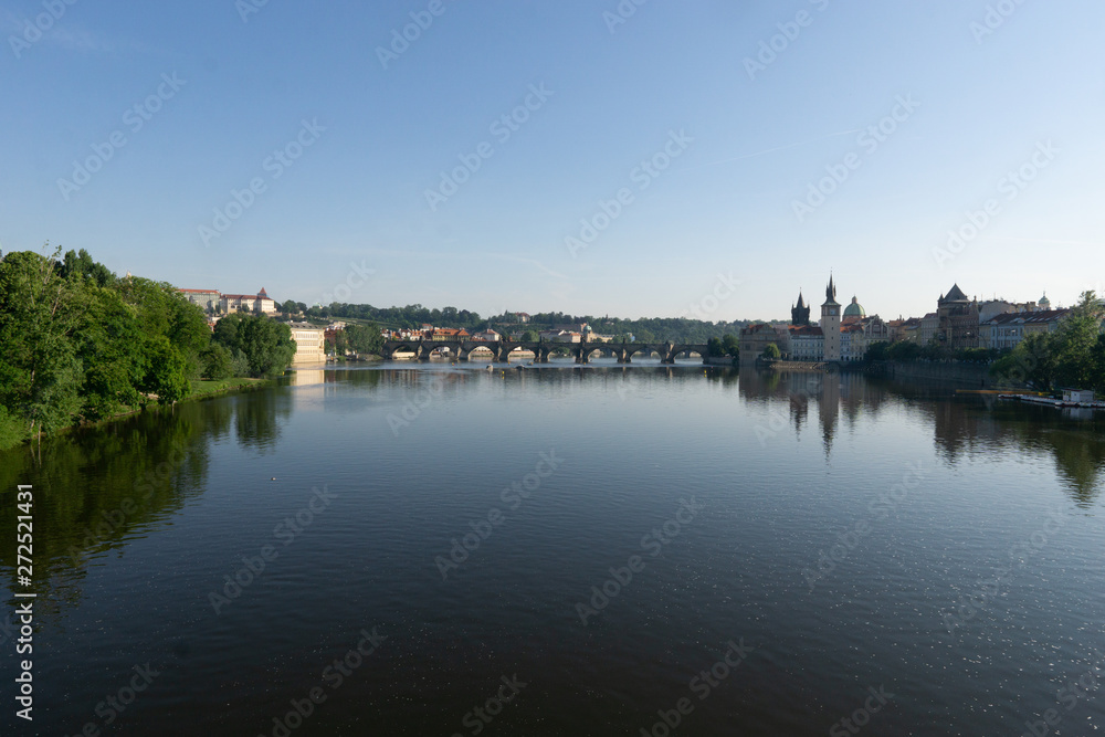 Charles Bridge is reflected in the calm surface of the water under the morning sun. The green foliage of trees.