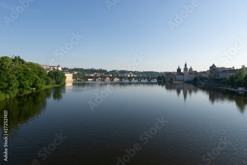 Charles Bridge is reflected in the calm surface of the water under the morning sun. The green foliage of trees.
